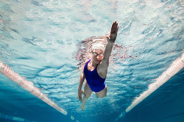 Mulher branca com maiô azul e touca branca em nado na piscina.