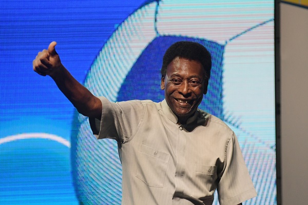 Pelé, homem negro idoso, dando um joia com a mão, ao fundo uma tela grande mostra uma bola de futebol