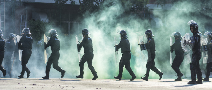 Policiais com escudos em meio à nuvem de fumaça simulando o decreto de um estado de exceção.