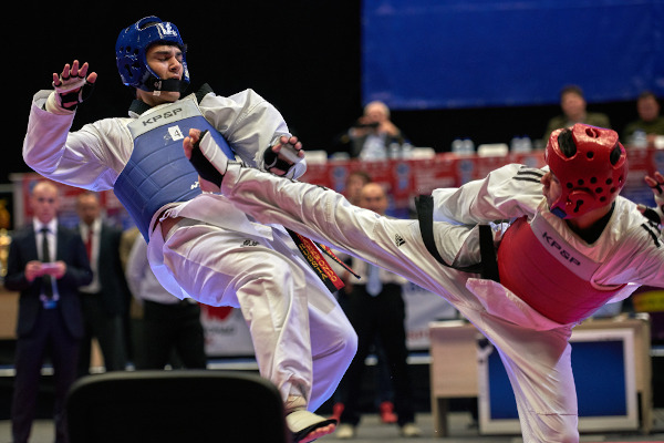 Atleta de taekwondo com protetores na cor vermelha realiza chute no adversário com protetores azuis.