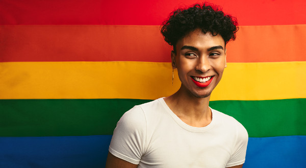 Retrato de uma pessoa queer e, ao fundo, a bandeira do orgulho LGBTQIA+.