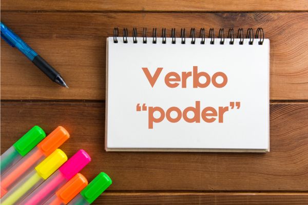 Canetas coloridas e lapiseira próximas a um caderno com o seguinte escrito: Verbo “poder”.