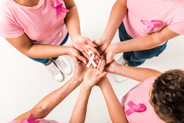 Mulheres de camisetas rosas e com laços rosas estendem as mãos. Ao centro, um laço rosa