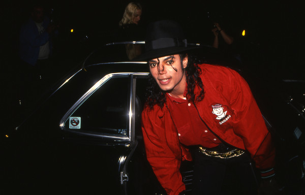 Michael Jackson de roupa vermelha e chapéu preto saindo de carro preto.