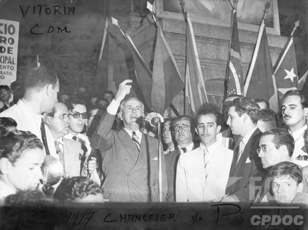 Oswaldo Aranha discursando em microfone e rodeado de homens.