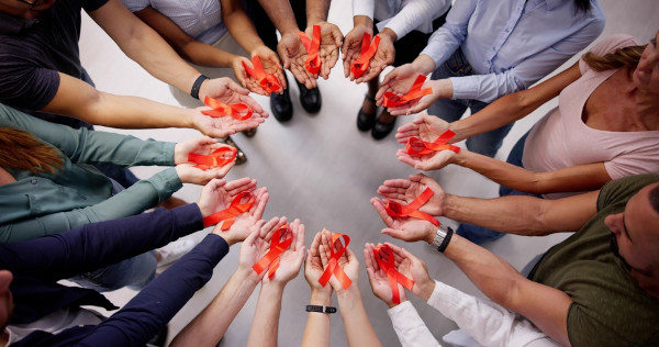 Várias pessoas, em círculo, com as mãos estendidas, cada uma com um laço vermelho, símbolo do Dezembro Vermelho.