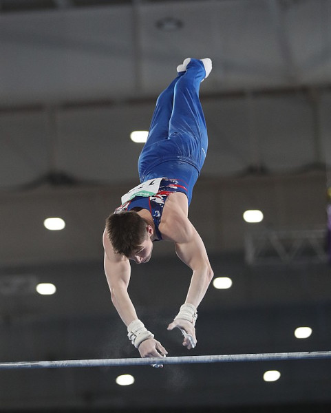 Competidor de azul realiza movimentação em barra fixa, modalidade de ginástica artística.