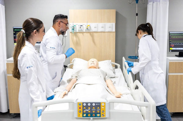 Professor ministrando aula à alunas da Faculdade Sírio-Libanês em um manequim deitado em leito hospitalar.