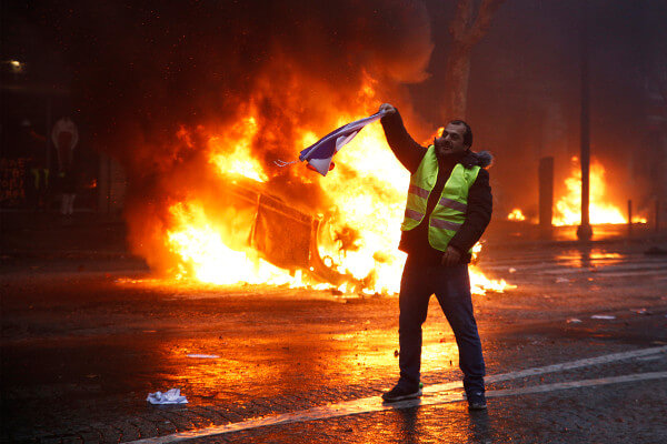 Homem usando um colete amarelo segura uma bandeira; atrás dele, um carro em chamas.